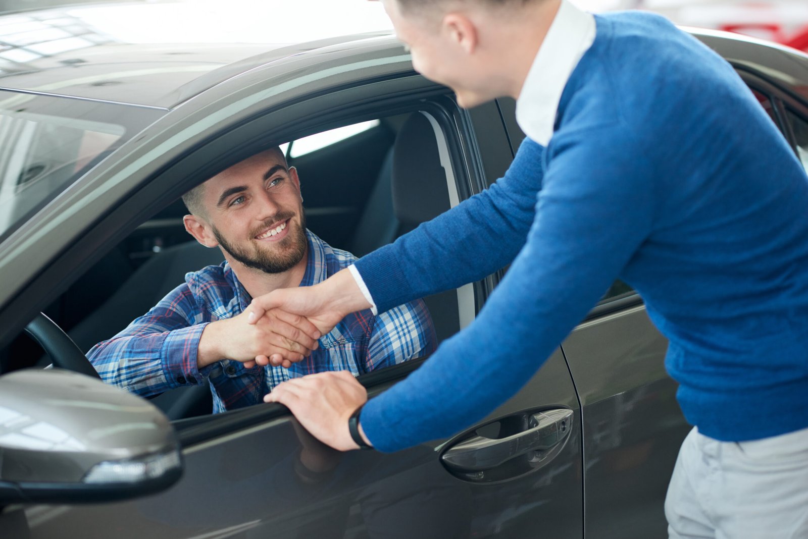 Unrecognizable male car seller and young bearded male customer handshake after new silver car purchase. Happy smiling handsome man in shirt sitting in car after successful vehicle buying.